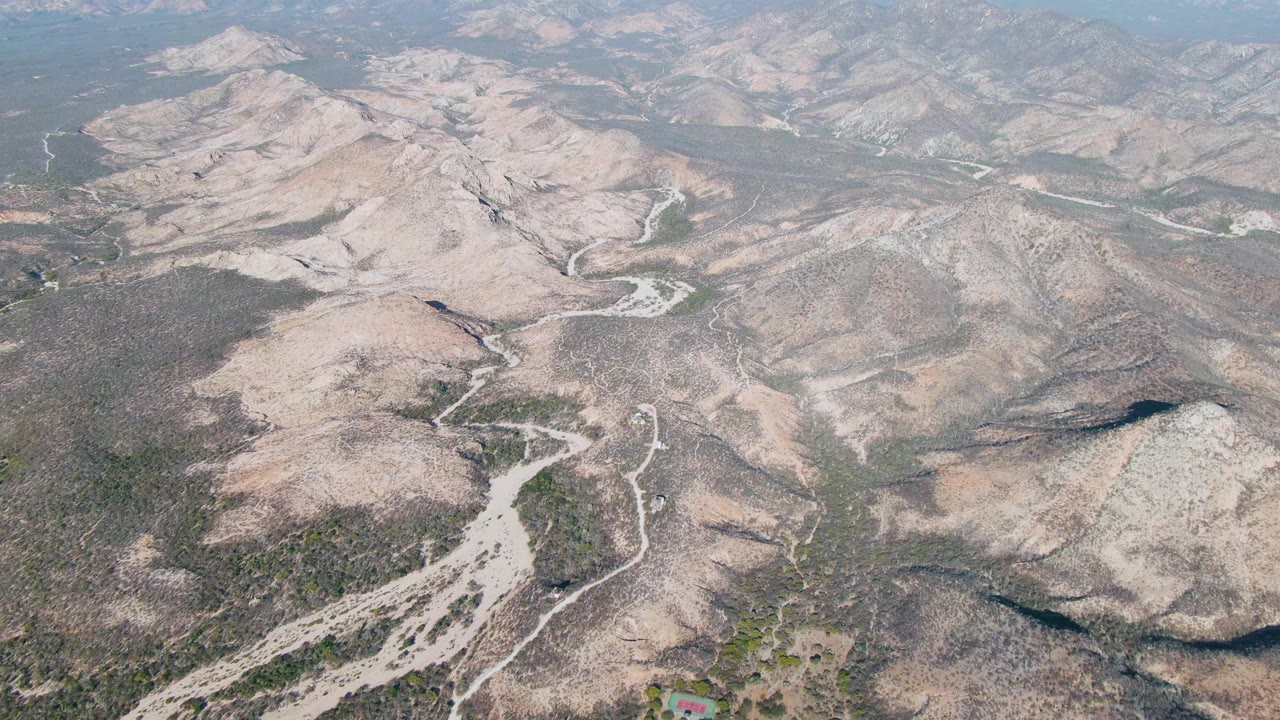 High aerial frame over Cabo Pulmo showing rocky beige hills, dark scrub patches, branching dry washes, a pale winding road, layered distant mountains, and no visible people.