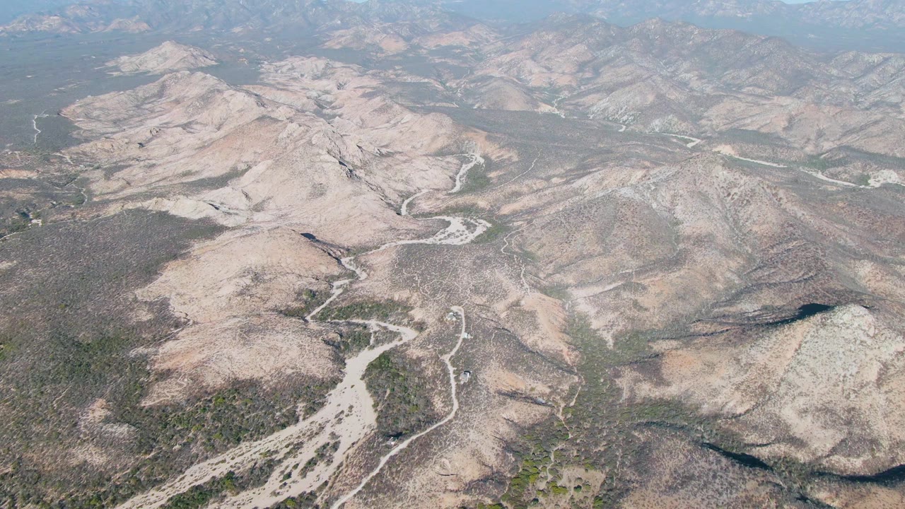 Drone-style aerial view in Cabo Pulmo with sunlit arid ridges, a curving dirt route through the midground, scrub-covered slopes, hazy mountain layers, clearing blue sky, and no visible people.