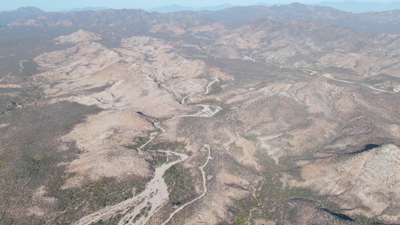 Opening aerial frame over Cabo Pulmo arid hills, pale dry riverbeds, sparse vegetation, distant ridges, and no visible people.