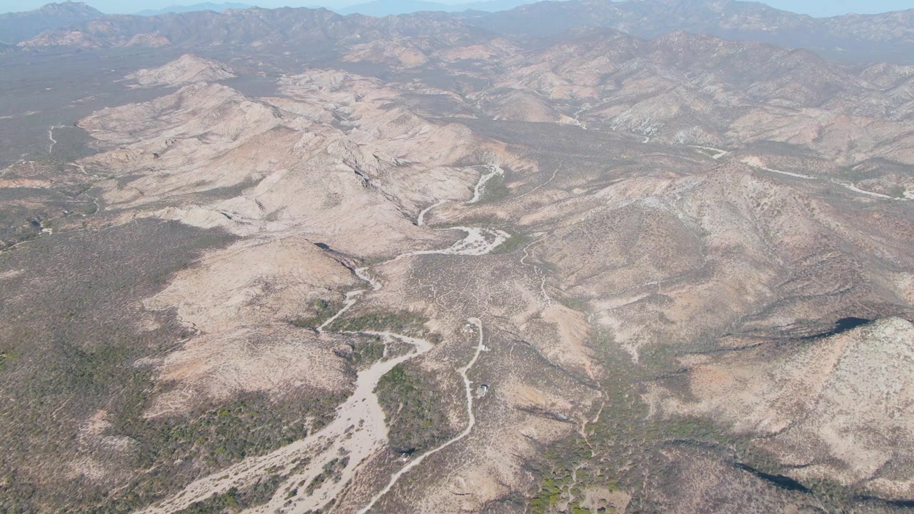 Aerial frame in Cabo Pulmo showing pale branching washes across tan hills, scrub vegetation in the low areas, a hazy horizon, and no visible people.