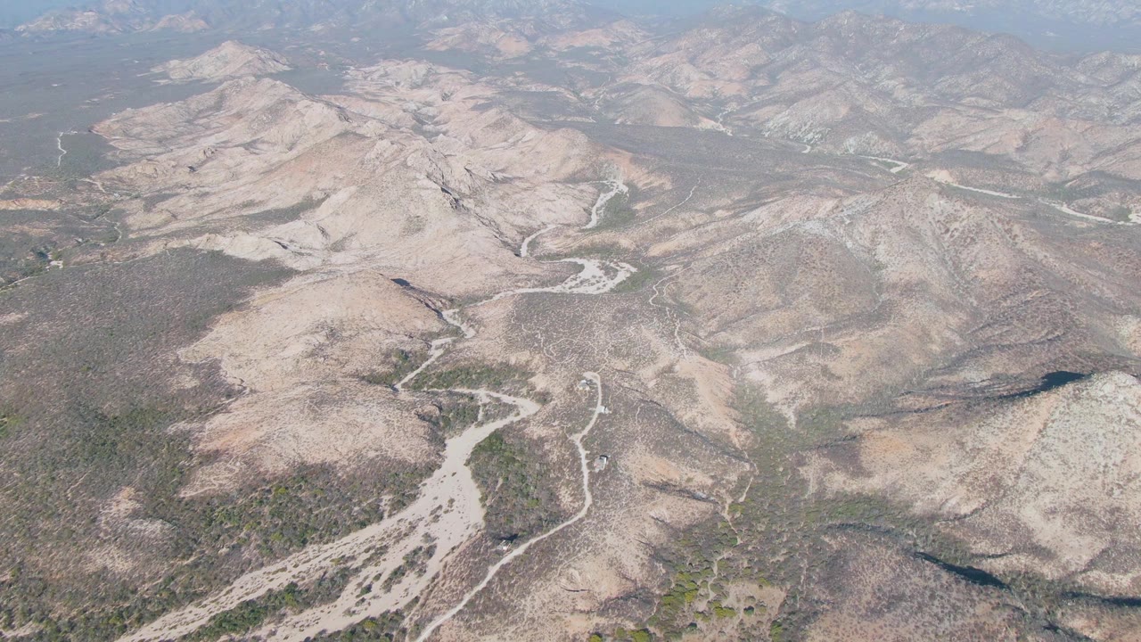 Mid-clip aerial frame over Cabo Pulmo terrain with winding dry channels, rocky slopes, scattered shrubs, distant mountain ridges, and no visible people.