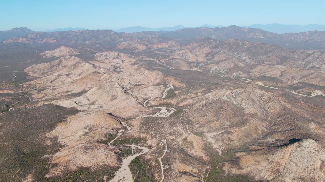 High aerial view near Cabo Pulmo, Baja California Sur, showing tan rocky hills, sparse scrub, a winding dirt road, distant mountains, and no visible people.