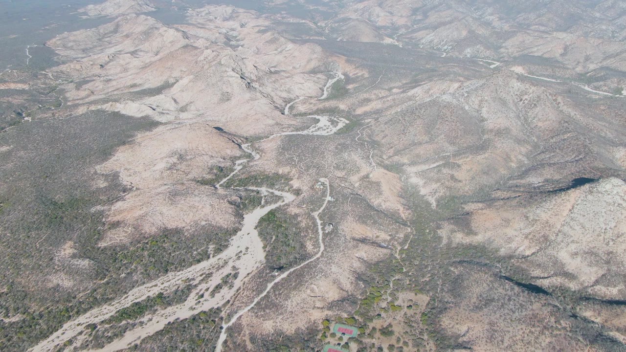 High aerial frame over Cabo Pulmo, Baja California Sur showing pale rocky hills, branching dirt roads, dry washes, sparse green vegetation, and no visible people.