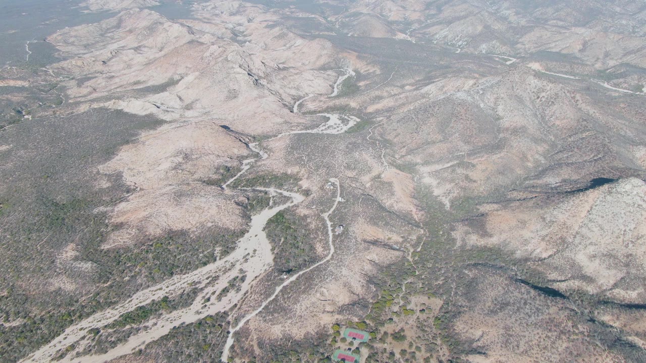 Aerial view in Cabo Pulmo, Baja California Sur with pale dirt tracks crossing scrub-covered slopes, dry drainage channels, layered hills, and no visible people.