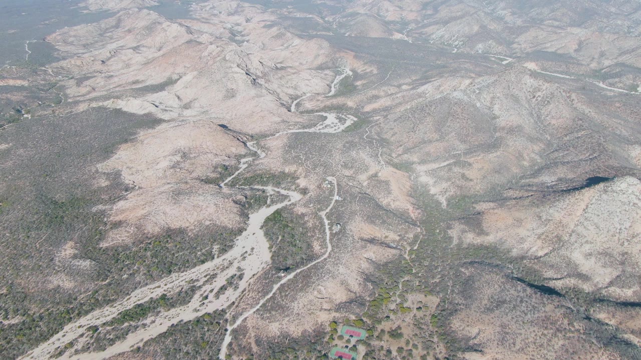Wide aerial frame over Cabo Pulmo, Baja California Sur showing branching dirt roads, tan and gray rocky hills, scattered green scrub, hazy background ridges, and no visible people.