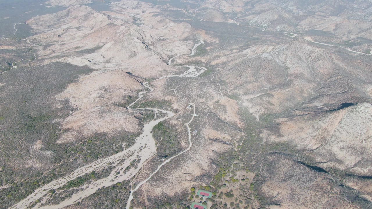 Aerial frame over Cabo Pulmo arid mountain terrain with pale branching dry riverbeds, sparse scrub, brown hills, a small red-roof structure near the lower edge, and no visible people.