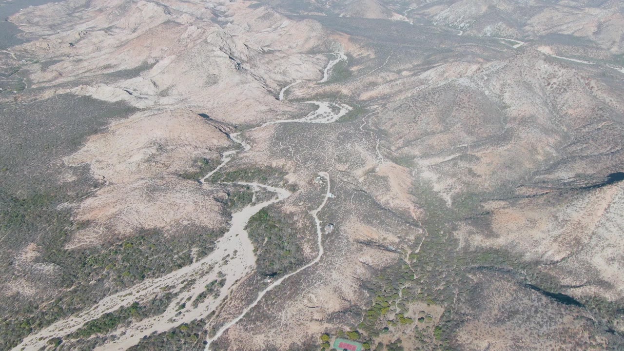 Aerial frame over Cabo Pulmo rocky beige hills with a bright dry riverbed branching through the lower center, narrow dirt paths on the slopes, and no visible people.