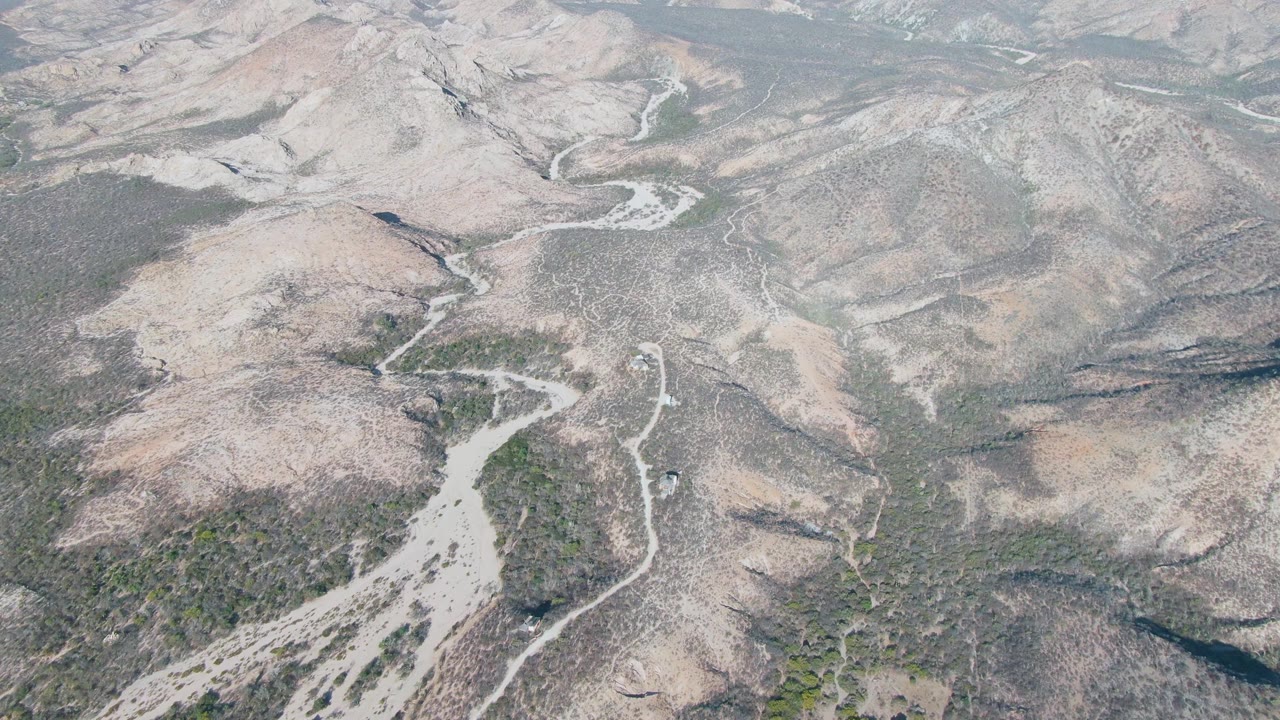 Aerial frame over Cabo Pulmo, Baja California Sur, with the pale stream channel slightly farther across the center, rocky slopes on both sides, scrub patches, and no visible people.
