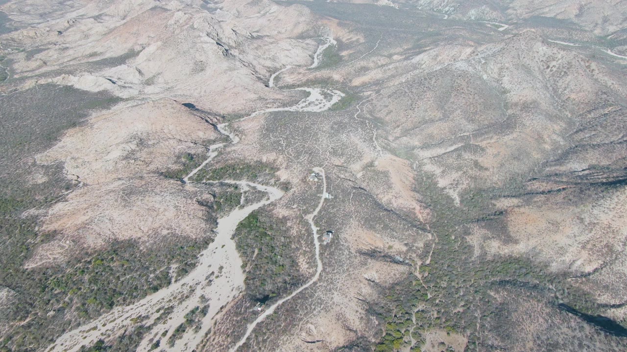Aerial frame over Cabo Pulmo, Baja California Sur, showing dry hills and a winding riverbed running from the lower area toward the upper terrain, with no visible people.