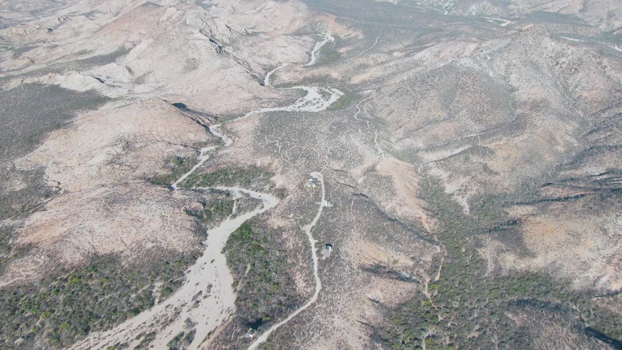 Aerial frame over Cabo Pulmo showing the dirt track bending near a pale dry wash, with scrub patches across rocky hills and no visible people.