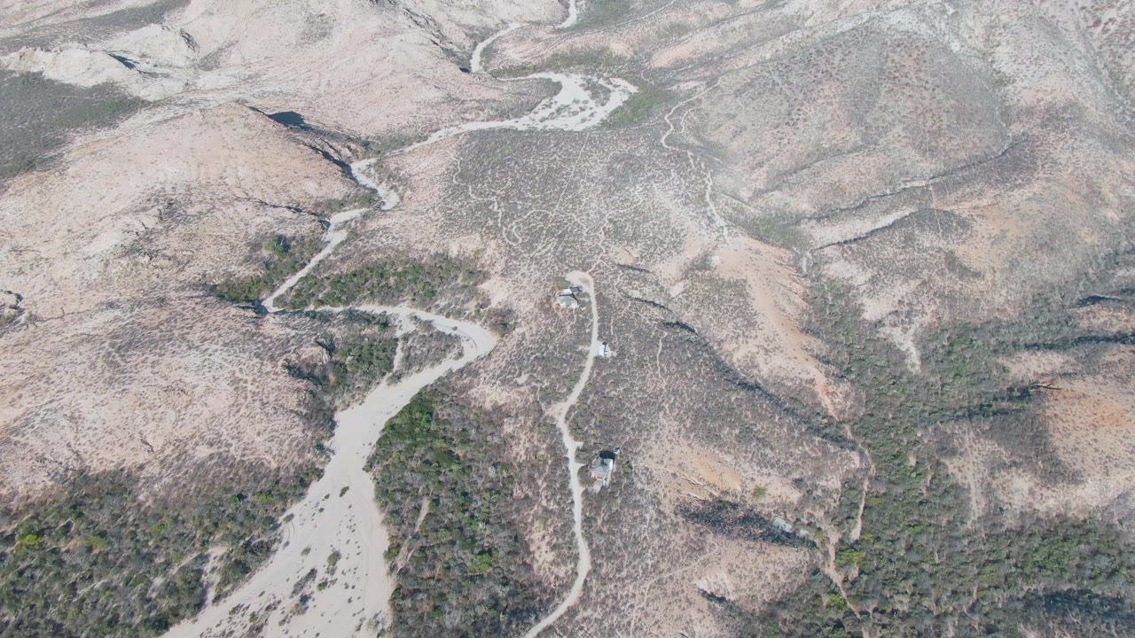 Drone view over Cabo Pulmo dry hills with the vehicle slightly farther along the curving dirt road, surrounded by pale washes and sparse green vegetation, with no visible people.