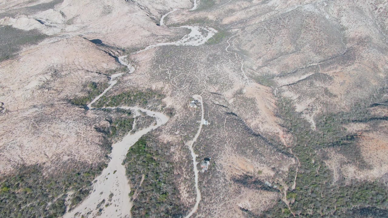 A high aerial frame in Cabo Pulmo with the small vehicle near a road bend, dry ridges filling the background and muted brown-green scrub across the terrain, with no visible people.