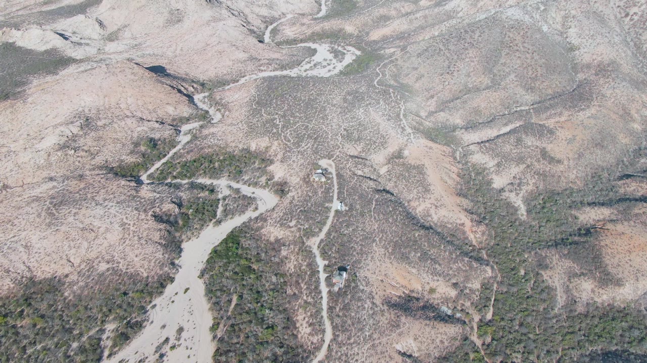 Wide aerial frame from Cabo Pulmo showing the dirt road snaking through a dry hillside valley, the vehicle near the lower road section, and no visible people.