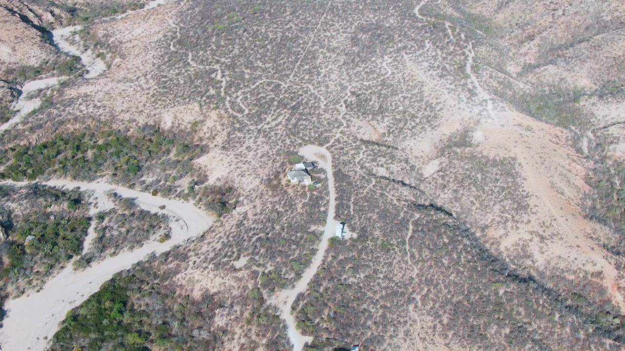 Aerial frame from Cabo Pulmo, Baja California Sur showing pale dirt roads crossing brown arid hills with sparse scrub, small roadside structures, a white vehicle near the road, and no visible people.