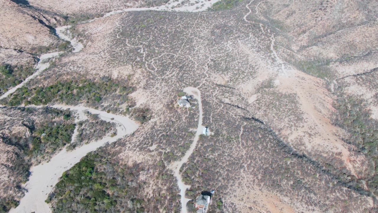 Aerial frame from Cabo Pulmo, Baja California Sur showing a pale road cutting through brown hills, a white vehicle on the road, scattered green scrub, small structures, and no visible people.