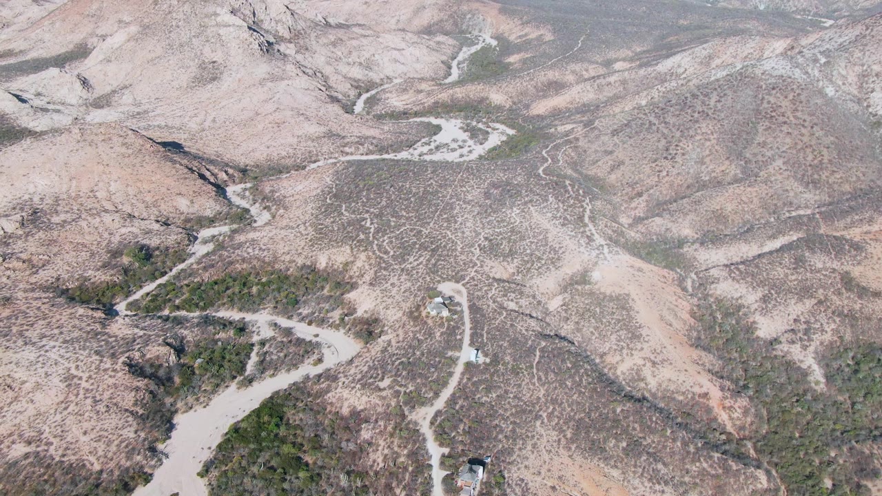 Aerial view over Cabo Pulmo dry mountain terrain with winding pale roads, sparse green vegetation, distant ridges, and no visible people.