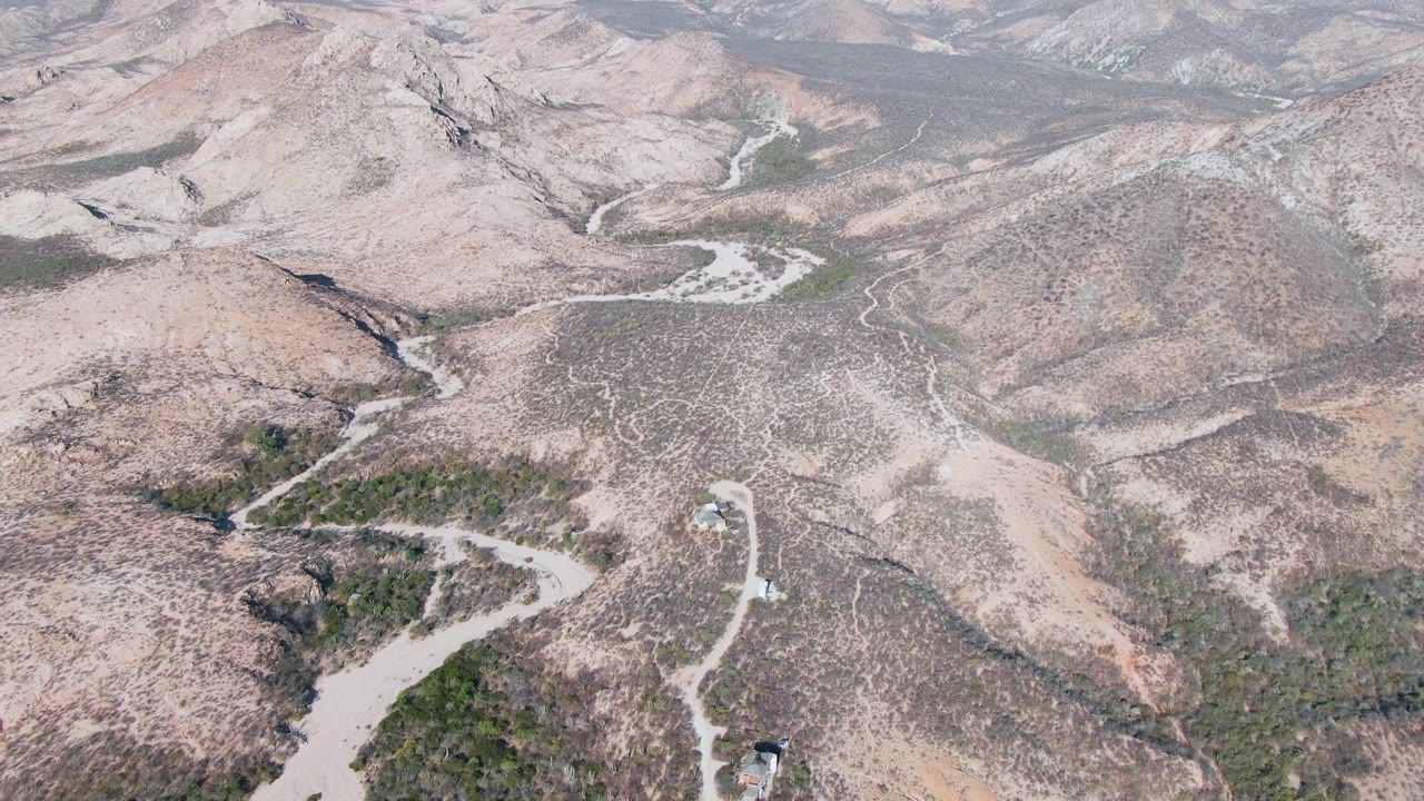 Aerial frame over Cabo Pulmo arid hills where a pale road curves through scrub-covered slopes toward layered background ridges, with no visible people.