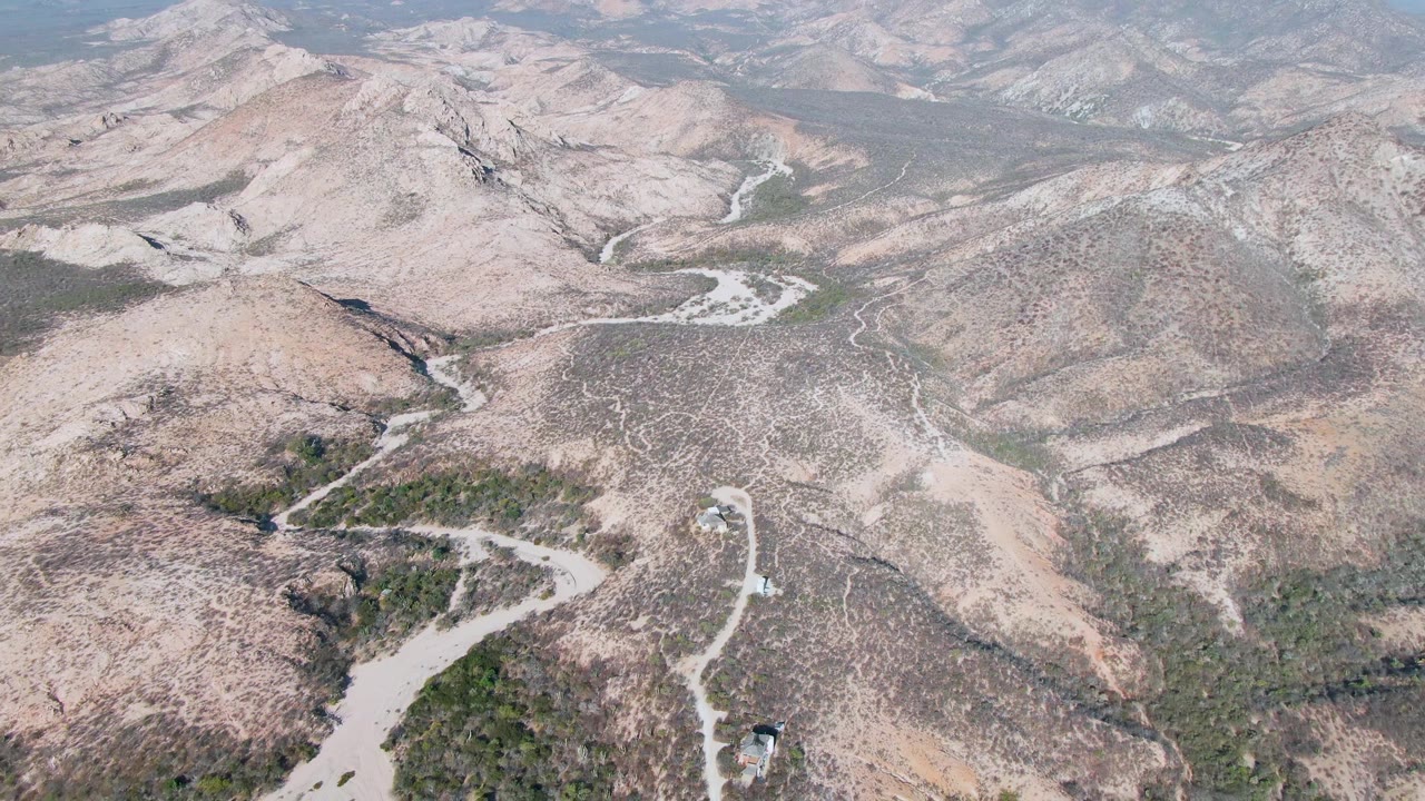 Aerial view in Cabo Pulmo showing branching pale roads, brown rocky hills, green scrub patches, and hazy mountains in the distance, with no visible people.