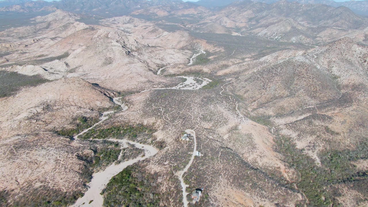 Aerial frame over Cabo Pulmo, Baja California Sur showing pale dirt roads crossing dry brown hills, scattered green vegetation, distant mountains, and no visible people.