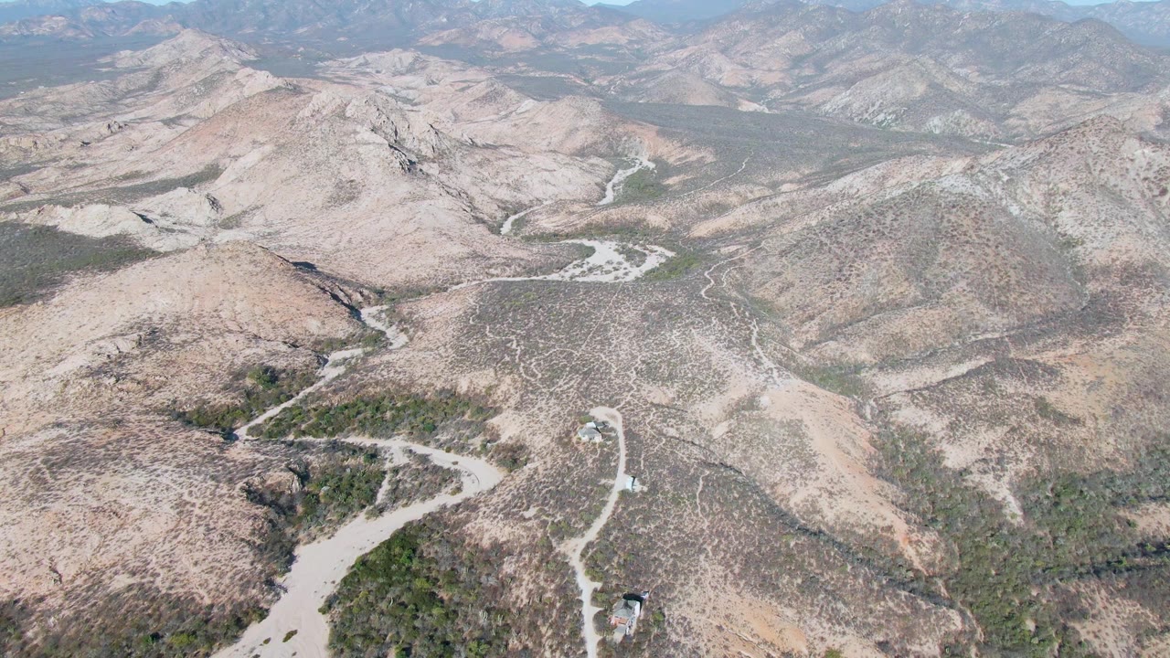 Drone-height view in Cabo Pulmo, Baja California Sur with a pale road bending around scrub-covered slopes, dry washes below, layered hills beyond, and no visible people.