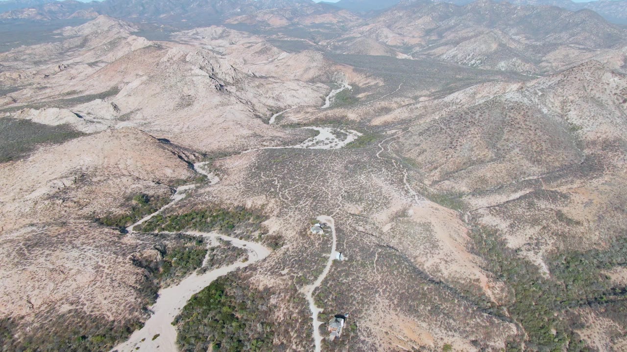 High aerial frame over Cabo Pulmo, Baja California Sur showing a winding dirt road, dry gullies, low green shrub patches, tan and gray hills, and no visible people.