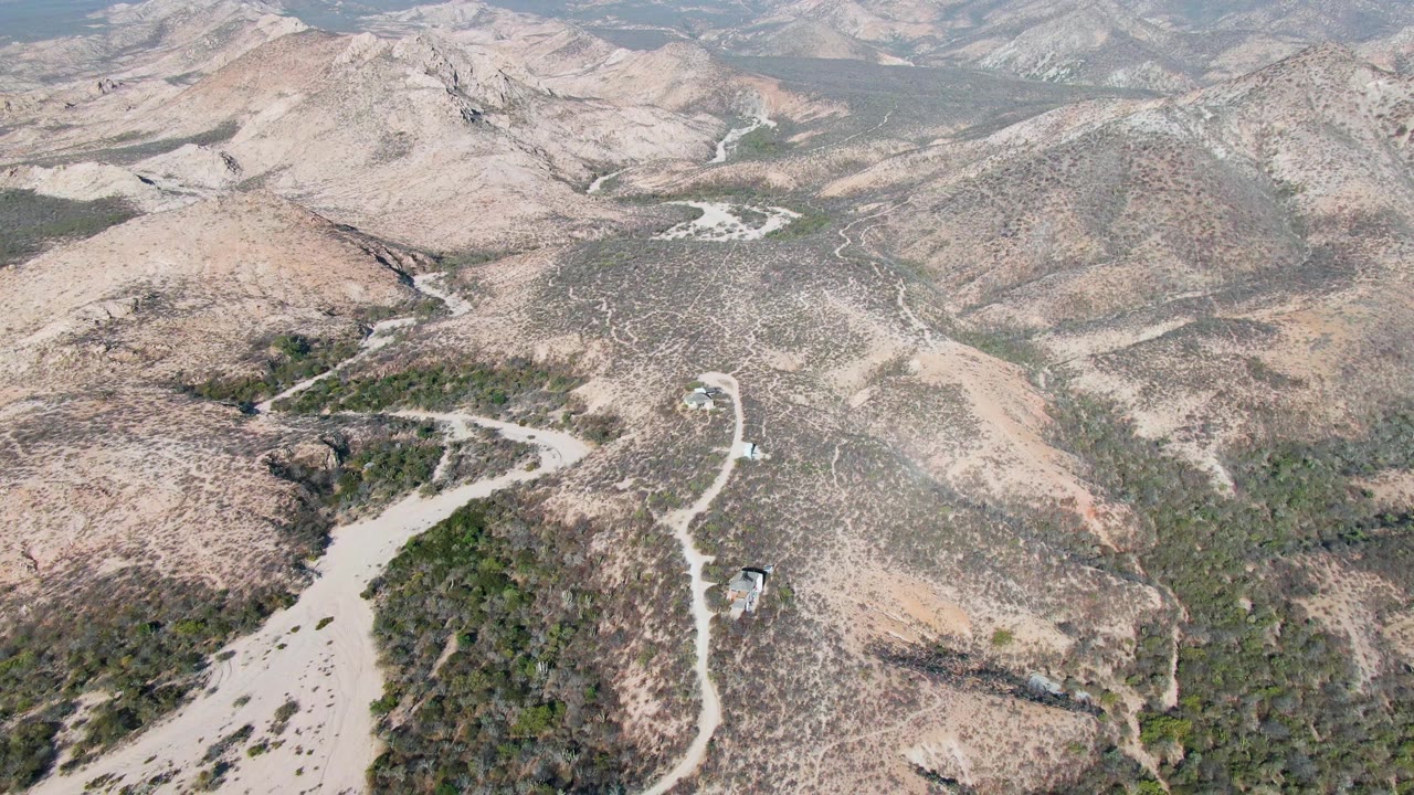 High drone view in Cabo Pulmo, Baja California Sur, with the vehicle slightly farther along the curving dirt road between tan hills and darker shrub patches, with no visible people.