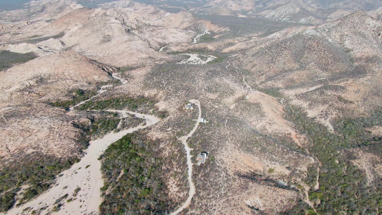 Aerial frame from Cabo Pulmo, Baja California Sur, showing the vehicle on the bright road near a bend, with dry slopes, scattered vegetation, and no visible people.
