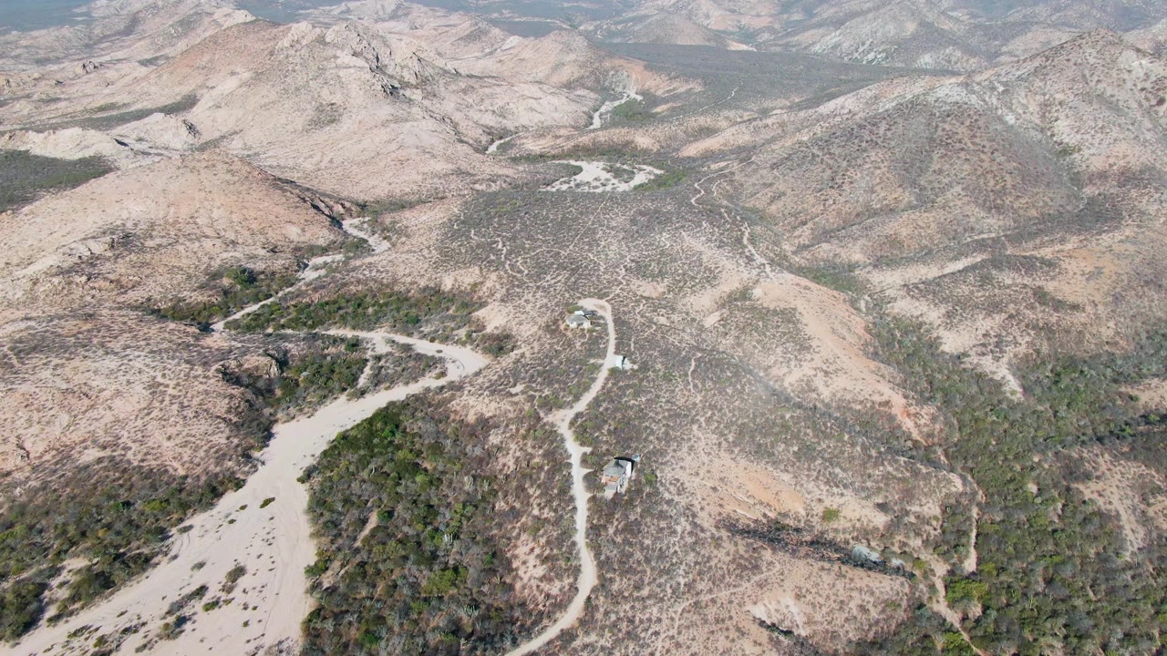 Drone view over Cabo Pulmo, Baja California Sur, with the winding road running from the lower frame into dry hills while the small vehicle remains visible and no people appear.
