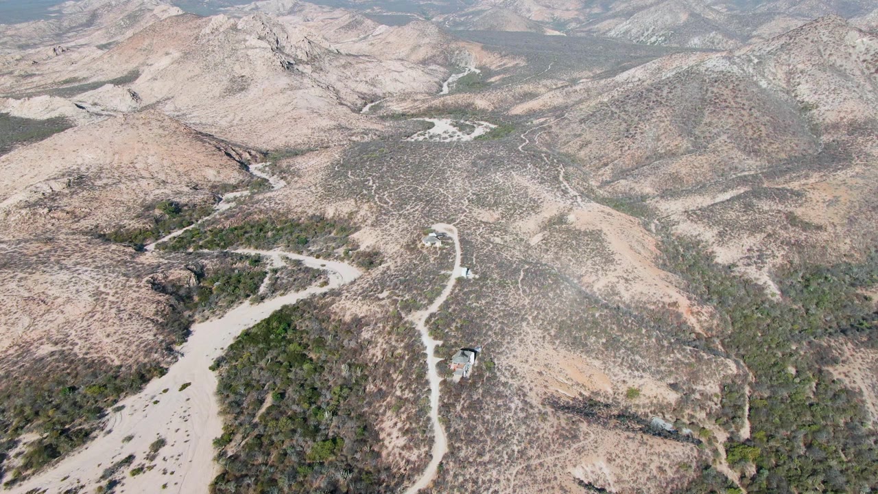 Aerial frame over Cabo Pulmo arid hills with winding pale dirt roads, rocky scrub slopes, distant mountains, hazy sky, and no visible people.