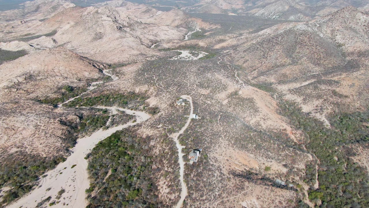 Drone-style view in Cabo Pulmo showing dry ridges, a pale dirt road curving through a wash, scattered scrub, distant mountains, and no visible people.