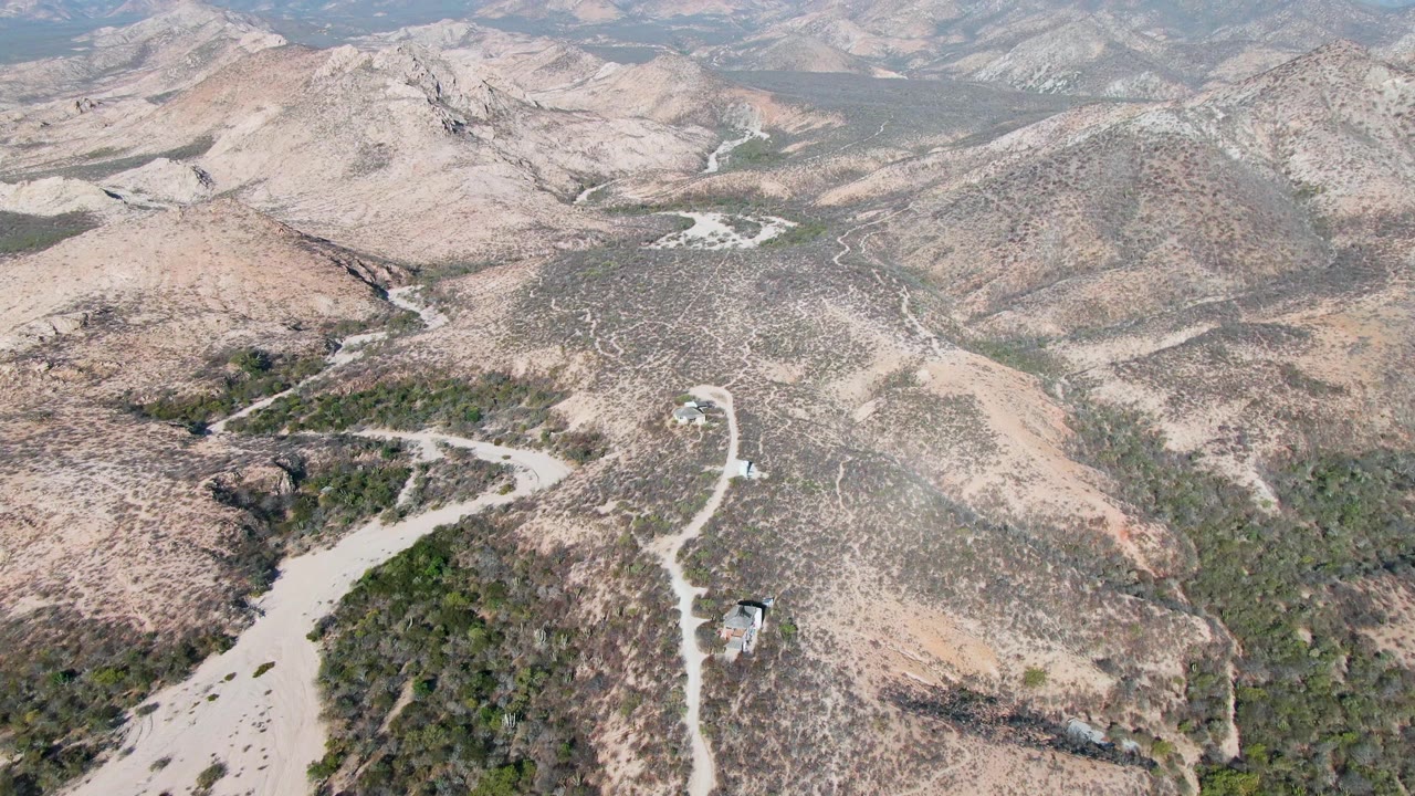 High aerial over Cabo Pulmo rocky desert hills with a winding dirt road, a small built element near the lower right road, distant ridgelines, and no visible people.