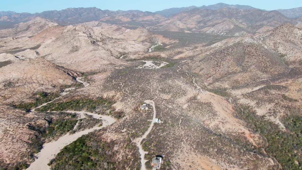 Aerial frame over Cabo Pulmo, Baja California Sur, showing a pale winding dirt road, a small vehicle near the lower road section, dry rocky hills, distant mountains, and no visible people.