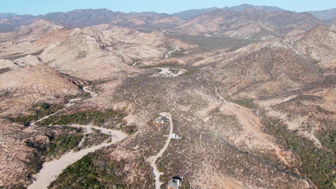 Drone view in Cabo Pulmo, Baja California Sur, with the road bending around a dry hillside, a small vehicle lower in frame, green patches in a wash, mountain ridges, and no visible people.