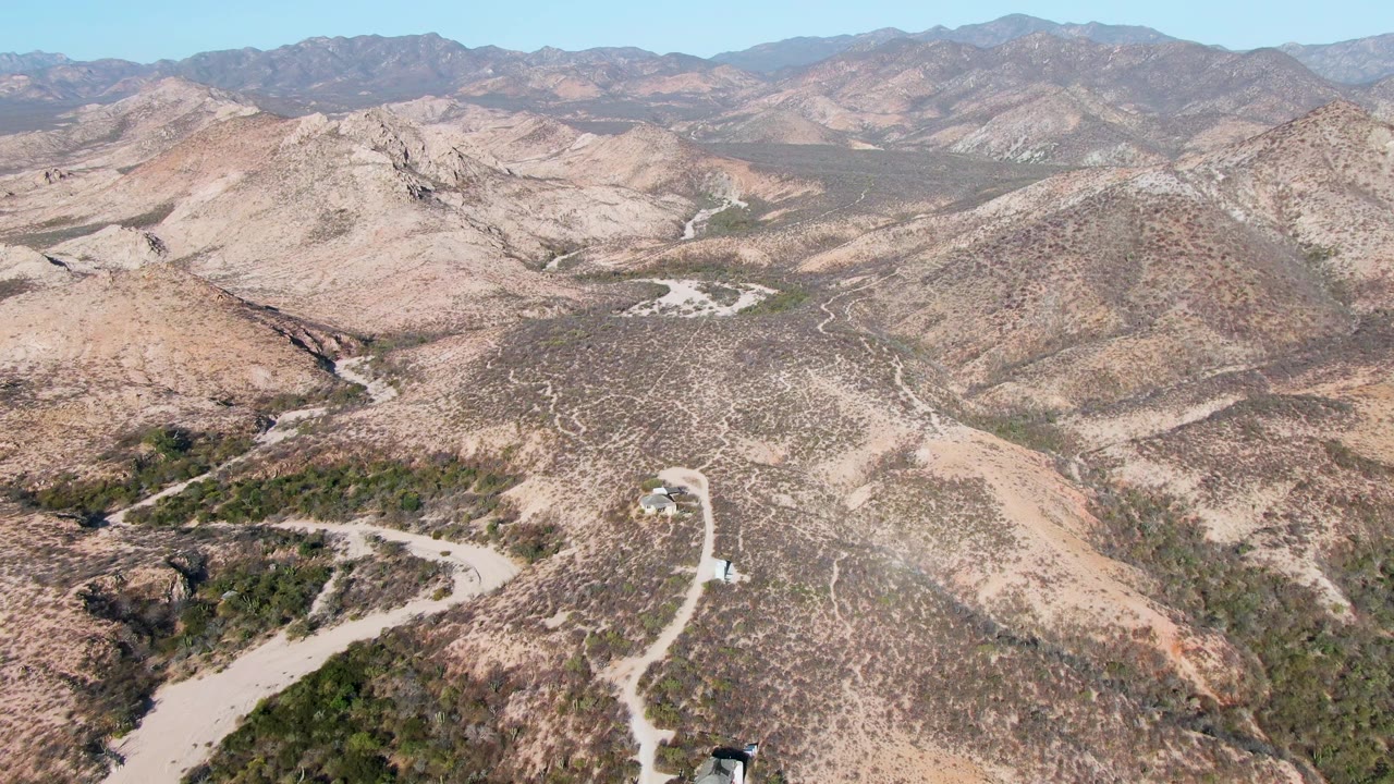 Wide aerial frame from Cabo Pulmo, Baja California Sur, looking across tan rocky hills, a curving dirt road with a small vehicle near the lower edge, layered mountains, and no visible people.