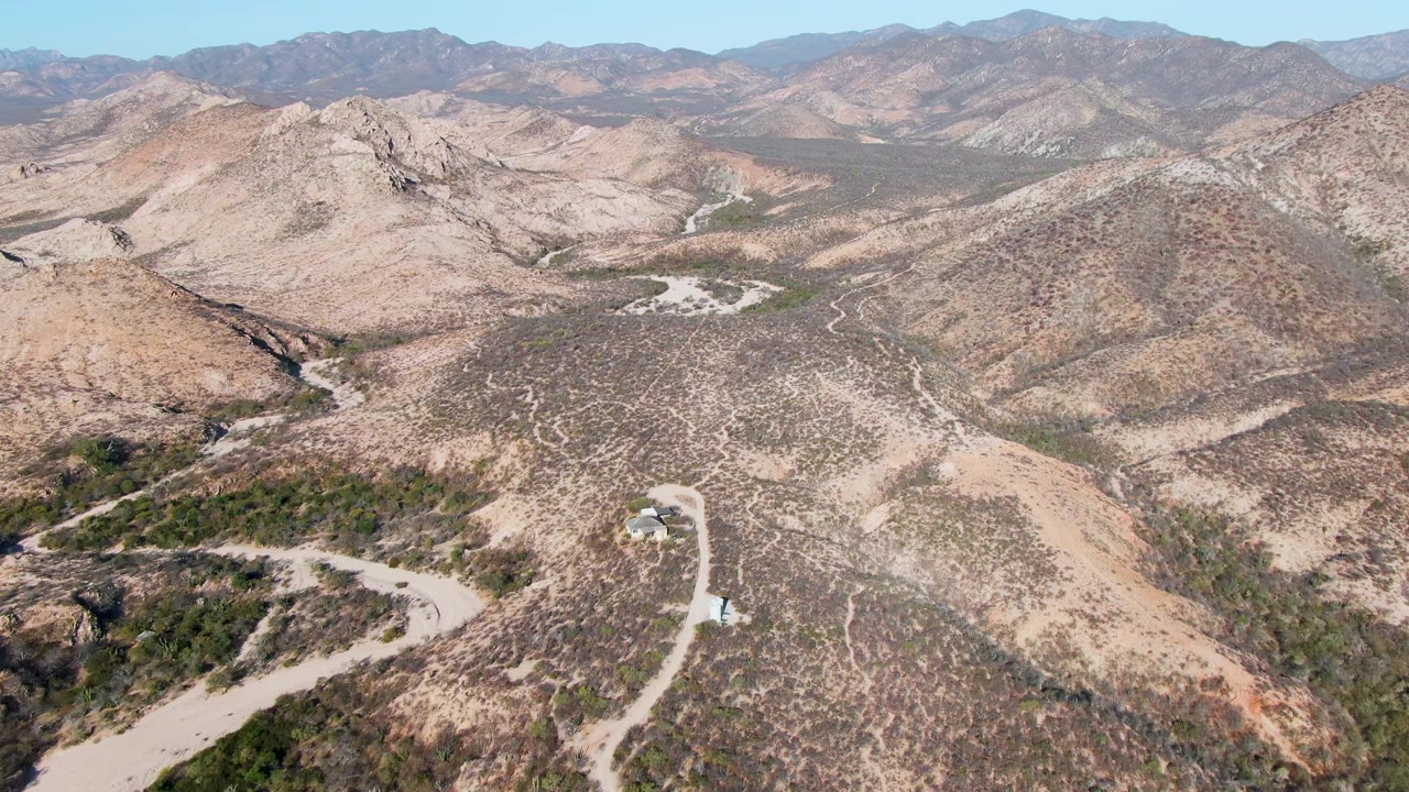Opening aerial frame over Cabo Pulmo desert hills, with a winding pale dirt road, sparse scrub, distant mountains, a small structure near the road, and no visible people.