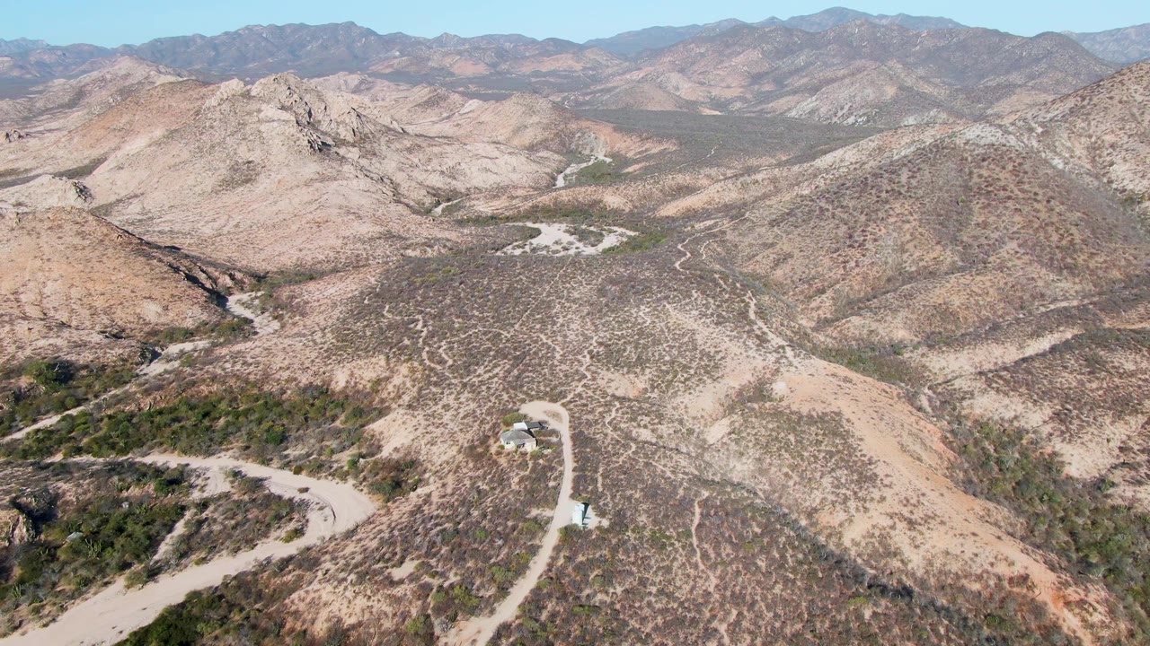 Mid-clip aerial view above Cabo Pulmo showing a pale road and wash lines cutting across dry hills, with a small vehicle or structure near the lower road area and no visible people.
