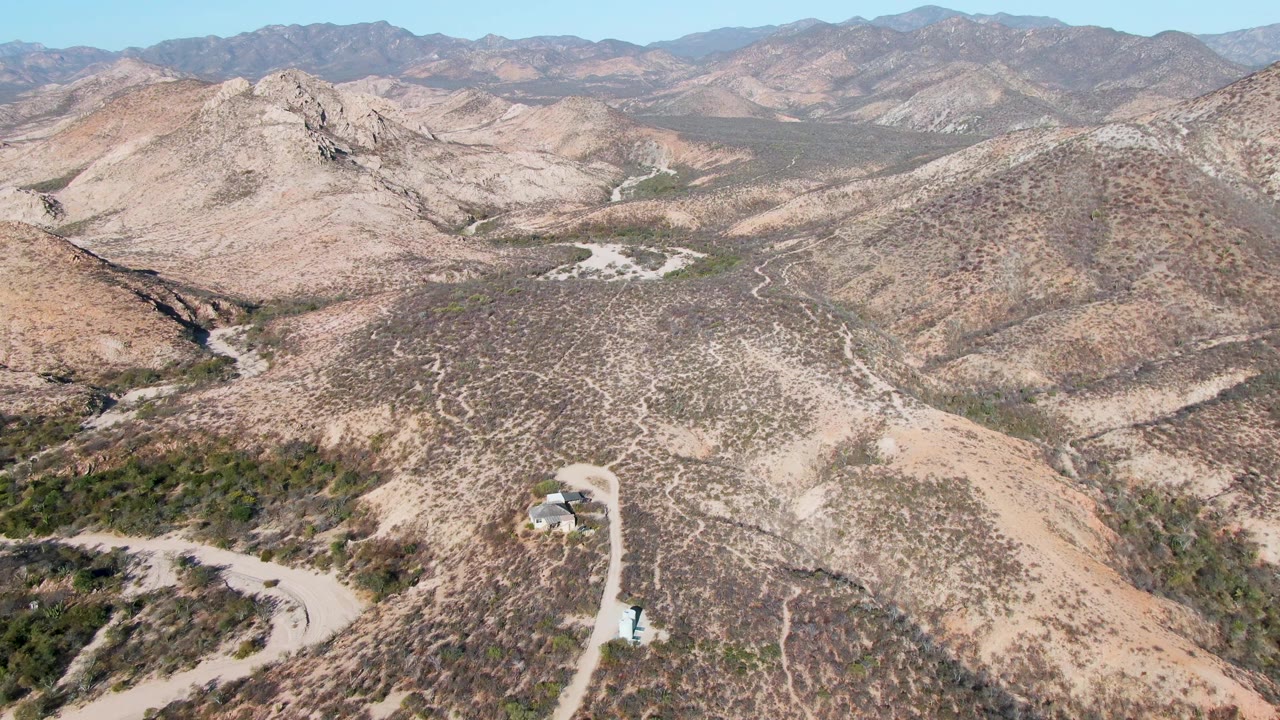 A high aerial view over Cabo Pulmo desert hills shows a pale winding dirt road, sparse scrub, distant mountain ridges, and no visible people.