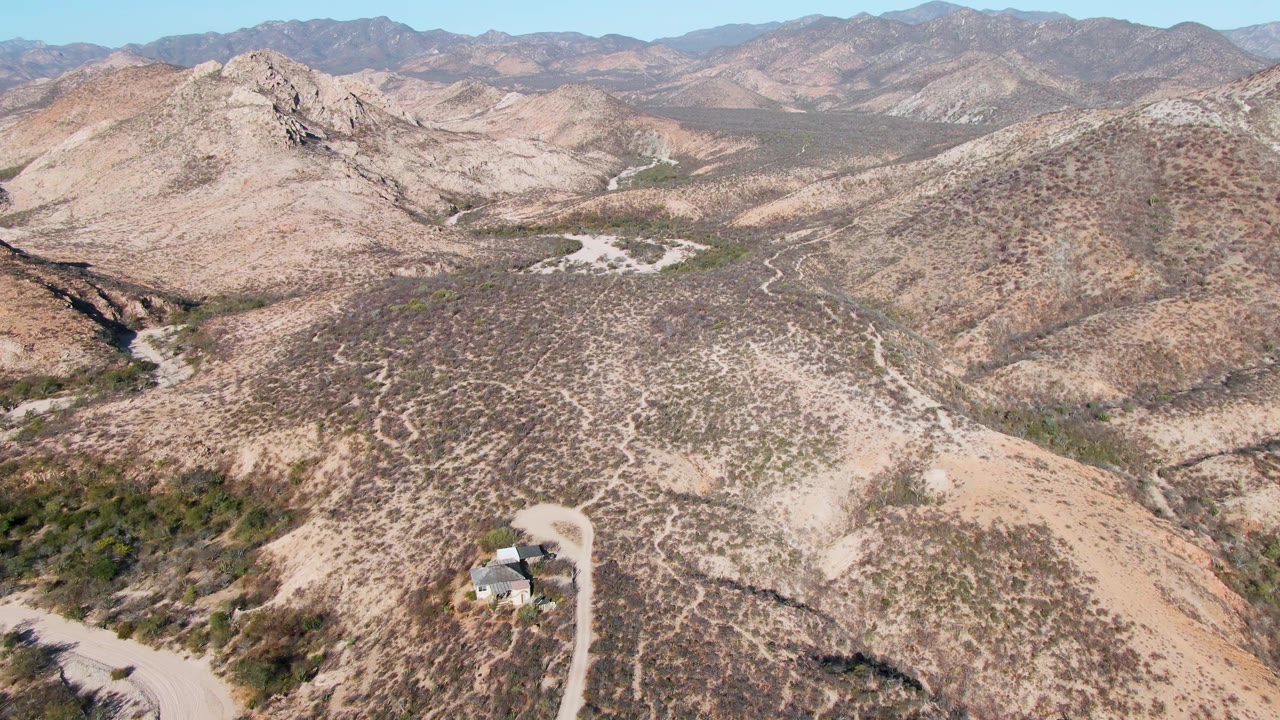 Opening aerial frame over Cabo Pulmo arid hills with a small building near the lower left, a dirt road, distant mountain ridges, clear blue sky, and no visible people.