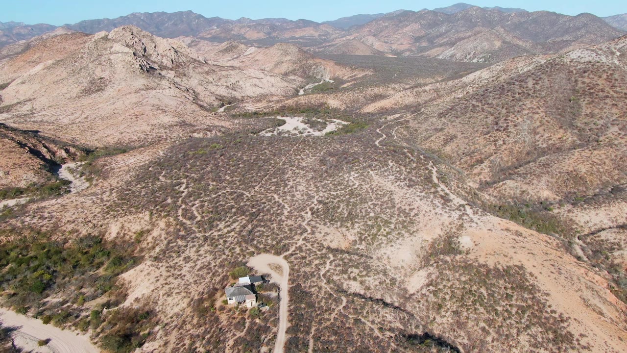 Aerial frame over Cabo Pulmo desert terrain with the small building lower in frame, pale dirt road curves, scrub-covered hills, mountain backdrop, and no visible people.