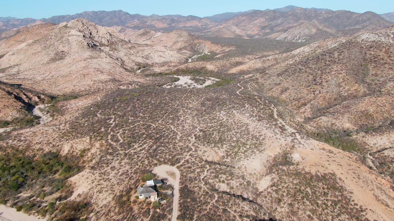 Mid-clip aerial frame in Cabo Pulmo showing scrubby tan slopes, a small structure by the road near the bottom, layered mountains beyond, clear sky, and no visible people.