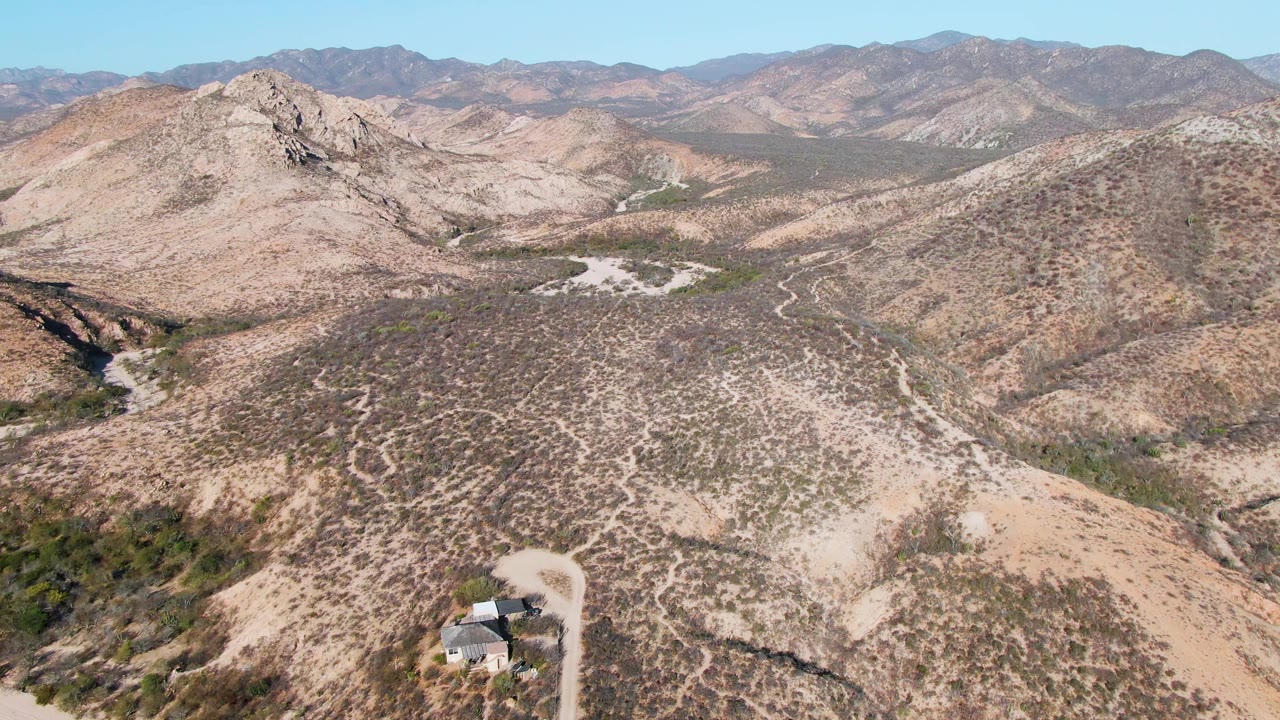 Wide aerial of desert ridges with a winding dirt road and small building near the foreground edge.