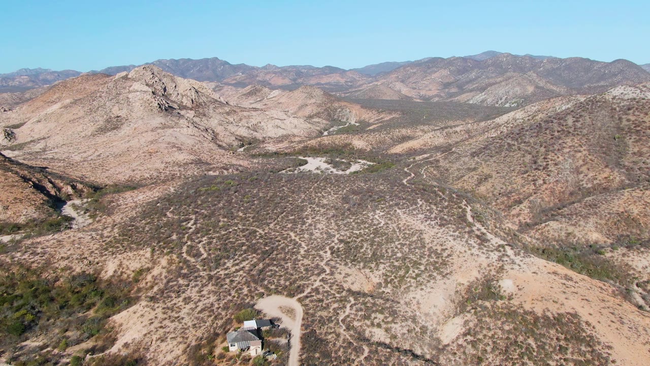 Aerial view over Cabo Pulmo desert hills with a small house near the lower left edge, a pale dirt road, distant mountain ridges, clear blue sky, and no visible people.