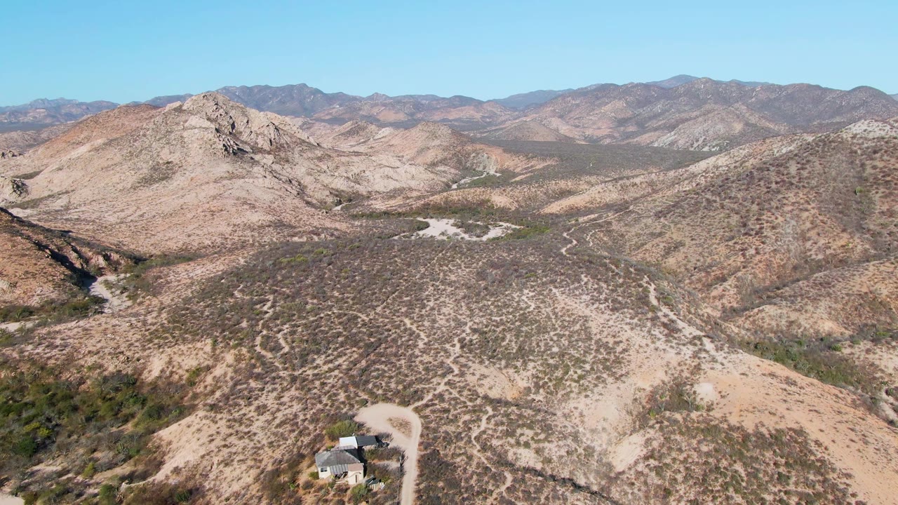 Cabo Pulmo desert valley seen from above with the isolated house lower in frame, scrub patterns across beige hills, a winding dirt road, mountain ridges, blue sky, and no visible people.