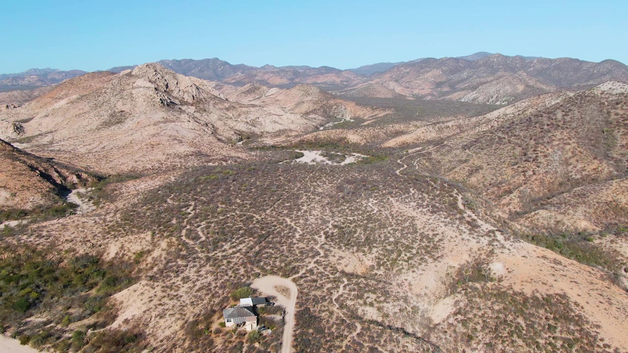 Aerial frame in Cabo Pulmo showing the house near the lower right, pale road curves below, brown desert hills in the midground, layered mountains on the horizon, and no visible people.