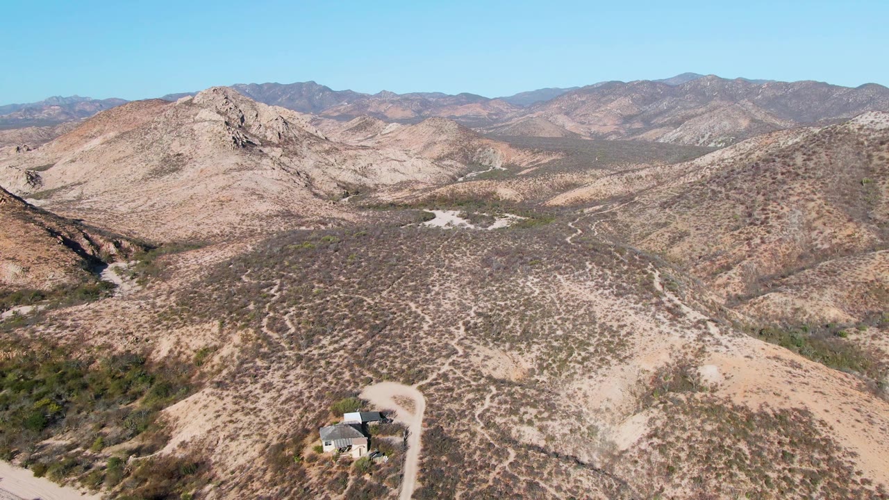 Wide aerial over dry hills with a house, dirt road, rocky ridge, and blue sky.