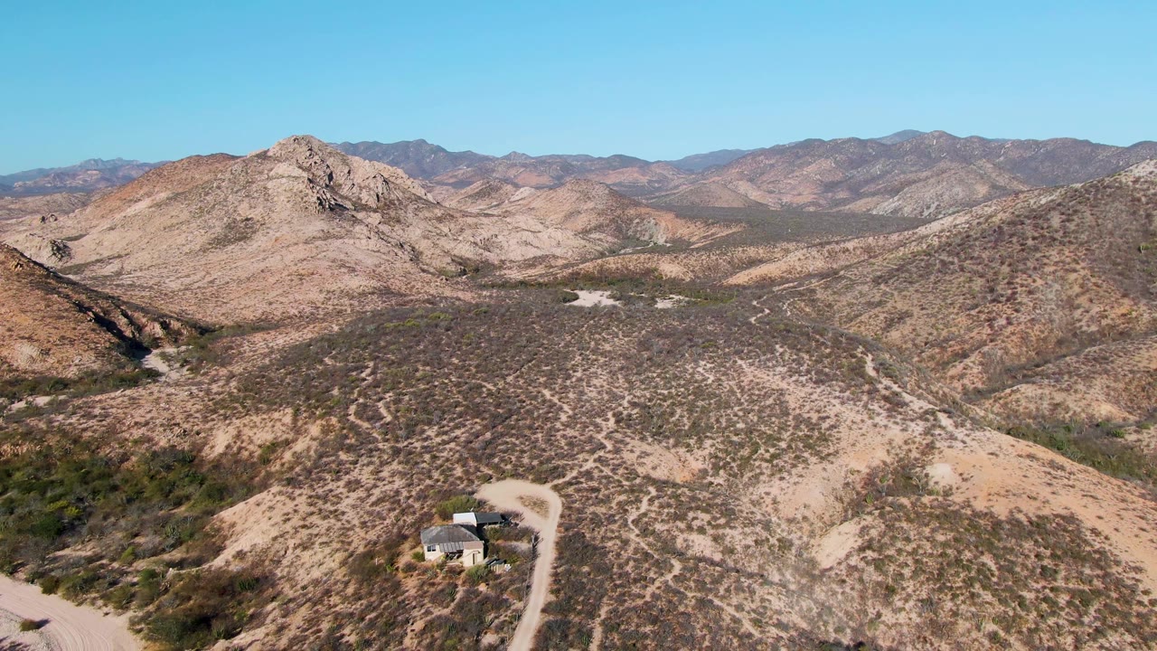 A small house and pale dirt road appear at the lower edge of a dry hillside in Cabo Pulmo, Baja California Sur, with rocky mountains and no visible people.
