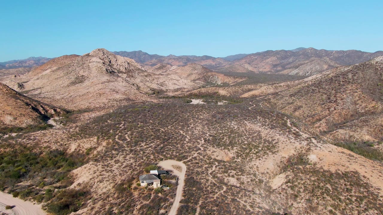 The house remains near the lower center of the Cabo Pulmo hillside frame, with a curved dirt road, scrubby slopes, layered mountains, and no visible people.