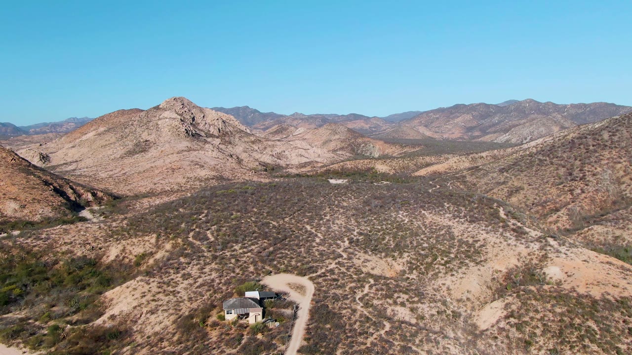 Wide aerial frame over Cabo Pulmo desert terrain, with a small house and winding dirt road at the lower left, scrub-covered hills in front, rocky mountains behind, and no visible people.