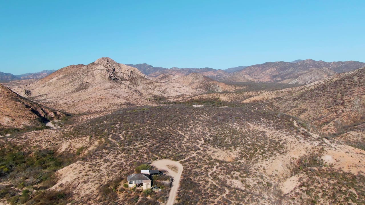 Drone-height view in Cabo Pulmo showing the small house slightly left of center near the lower edge, a pale dirt road curving beside it, dry scrub slopes, distant ridges, and no visible people.