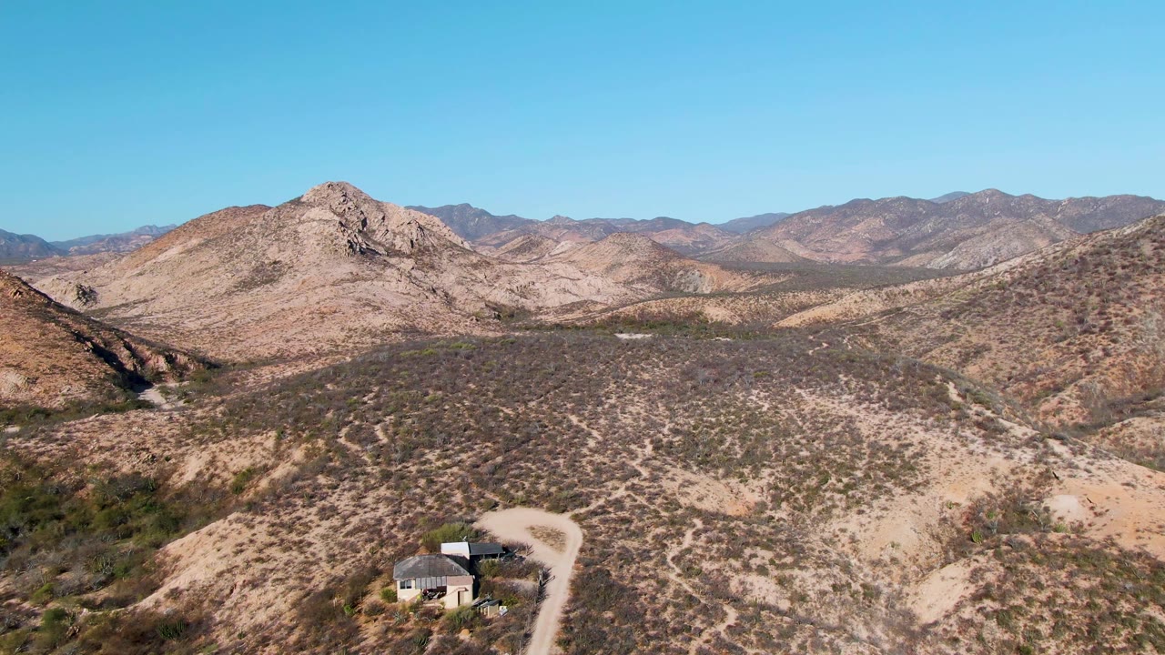 Aerial frame from Cabo Pulmo with the house near the lower middle, a dirt road bending around it, tan desert slopes in the midground, layered mountains in the distance, and no visible people.